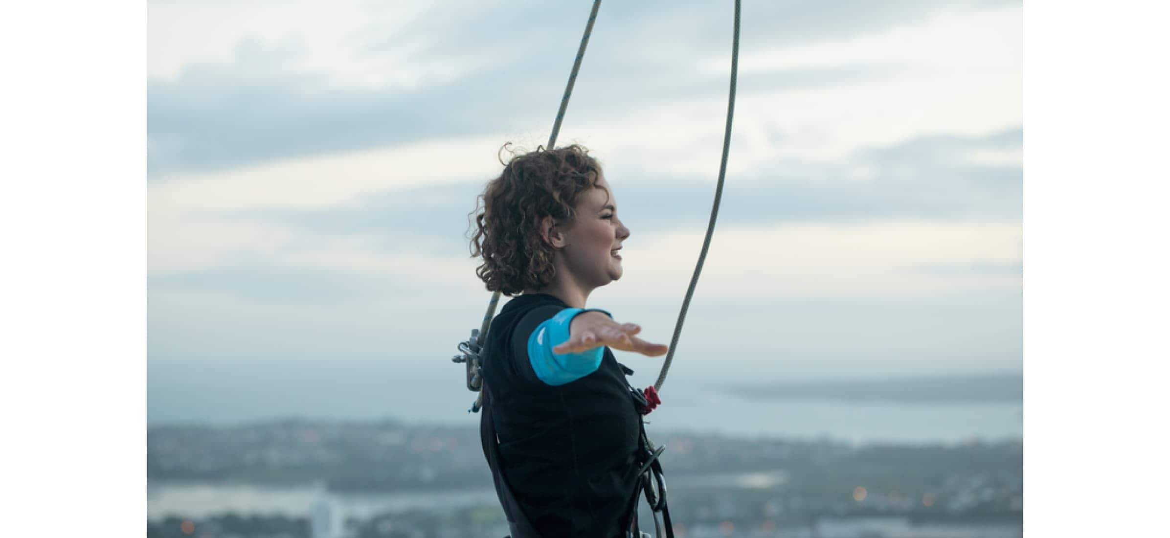 Phoebe Heyhoe standing with her arms outstretched wearing a vest with ropes attached to it, standing somewhere high up.